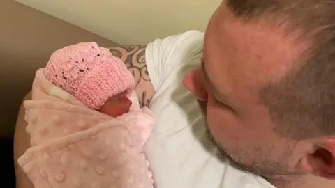 Family handout A tiny premature baby wrapped in a pink blanket and wearing a pink beanie is held by a man.