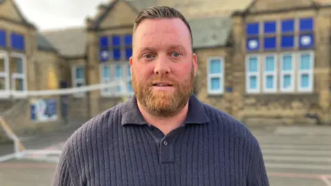Kevin Precious stands in a school playground looking at the camera. He has short brown hair and a reddish-brown beard. He is wearing a grey knitted top. Behind him is a football goal in front of a row of stone school buildings. 