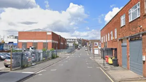 A long street with a car park on the left as well as large red brick buildings surrounded by metal fencing. On the right are red brick buildings with grey metal shutters over garages 