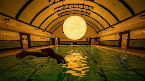 A man dives into a dimly lit swimming pool. Above the pool, suspending from an arched ceiling is a large glowing sun.