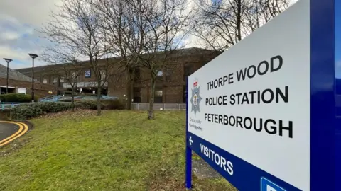 A sign reading "THORPE WOOD POLICE STATION PETERBOROUGH" on a grass verge outside a two-storey brick building.
