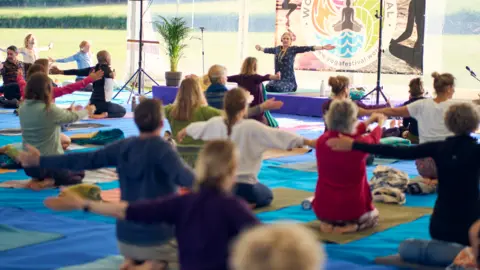 People in a marquee doing yoga and taking instructions from a teacher on a stage.