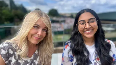 Carolle Forde Garcia and Iman Mahmood are in a stand at Wimbledon with the courts and the crowds behind them. They both have long hair and are smiling.