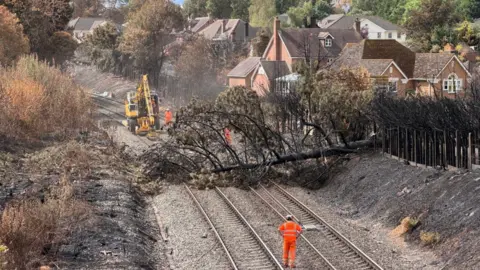 Network Rail A train fallen across a railway line, which has scorced land on either side of the tracks, as workers wearing orange hi-vis walk on the tracks, with a yellow digger seen behind.