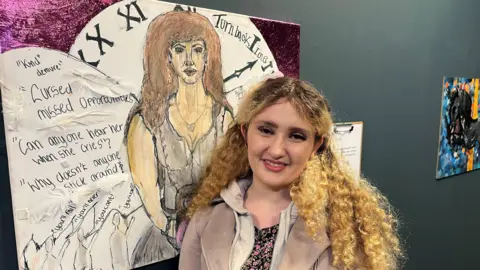 Federica Bedendo/BBC Kimberley Jackson with her artwork. She has long blond curly hair and dark eyes. She is wearing a black and pink flowery blouse with a cream jacket over it. Her artwork shows a big clock and words such as "can anyone hear her when she cries". In front of the clock is a female figure with wings.