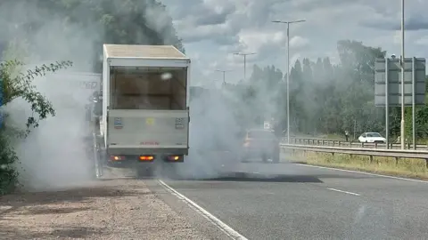 BCH Road Policing Unit A white box van, on a dual carriageway, with lots of smoke all around it. Other cars are on the road, and a white car, driving in the opposite direction. There are trees around the road and a large road sign. 