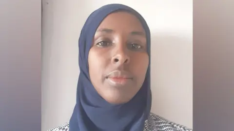 Fadumo Haji A black woman in a blue headscarf posing for a picture. The background is blurred. She is against a clear wall with a slight shadow. She is wearing a black and white winter coat. 
