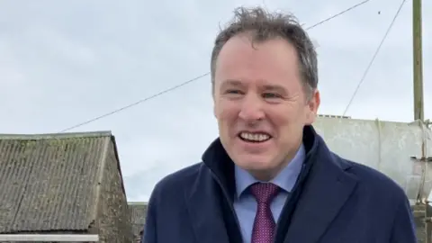 Shows a man smiling in a blue shirt, purple, navy blue overcoat and standing in front of telegraph pole with cloudy sky in the background