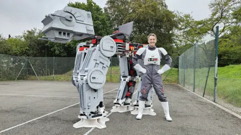 Jame Bruton James Bruton dressed in something resembling a Star Wars Stormtrooper outfit, stands next to his homemade At-At walker.