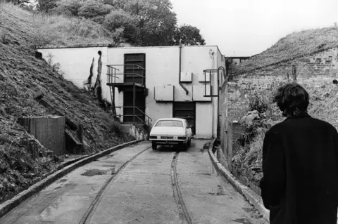 Media Wales Black-and-white photograph of a concrete service building set into a hillside, resembling a bunker or underground facility entrance. A narrow roadway with rail tracks embedded in the surface leads to the building, where a small car is parked outside. In the foreground, a person wearing a dark coat is seen from behind looking toward the structure. Steep grassy banks and retaining walls flank the entrance, creating a secluded, industrial setting.