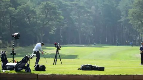 Getty Images A man hitting a golf ball with a golf club down a large patch of grass lined with tall trees. There is a TV camera filming him 