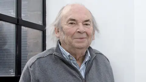 Quentin Blake with long grey hair wearing a grey jumper and blue and white striped shirt standing in a building with white painted walls in front of a window.