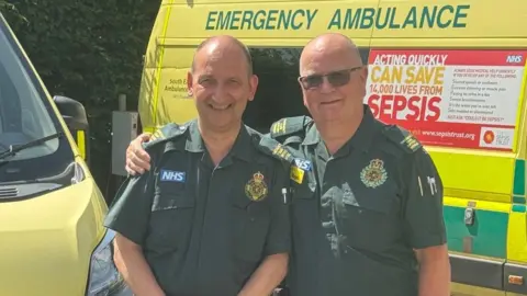 Two men in green paramedic uniforms stand in front of an ambulance in the sunshine. The man on the right has his arm around the other's shoulder.