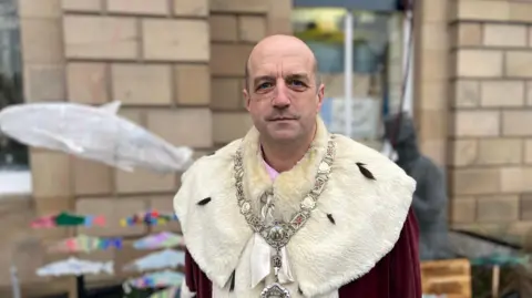 A bald-headed man wearing a large silver ceremonial chain and robes looks at the camera