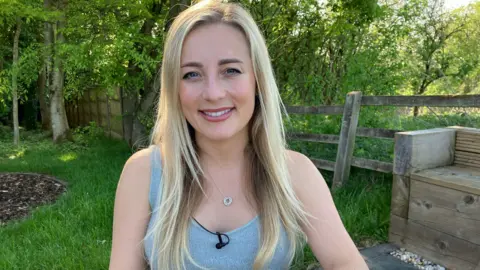 Eve Morgan, blonde and smiling at the cameras earring a grey vest and small pendant necklace. She is sitting in a garden area with grass and trees behind her