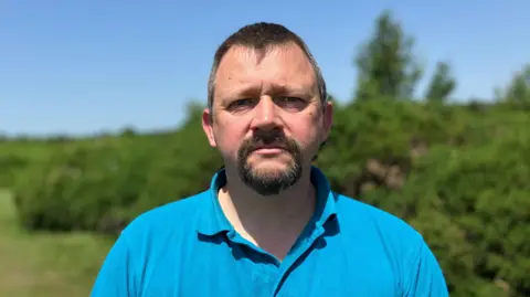Simon Lee stands outside in front of green hedges and with a blue sky above. He is short‑haired and bearded, and is wearing a turquoise polo shirt.