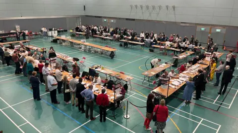 BBC A sports hall full of people standing one side of long tables opposite people sitting down with papers spread out in front of them.