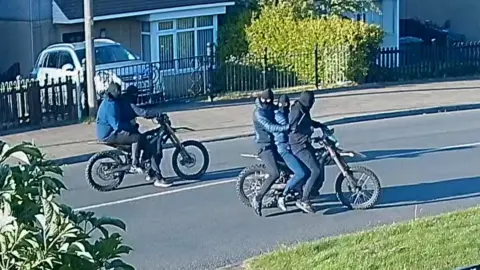 Two masked people ride one motorbike, while three masked people ride another motorbike nearby. All five of them are wearing dark jackets, jeans and balaclavas. 