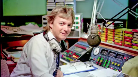 Pete Baker, de Piccadilly Radio, se sienta detrás de la mesa de mezclas con una camisa blanca y auriculares colgados del cuello.