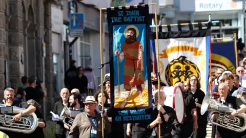 Penzance Council People through a town centre. Some are carrying brass instruments. Other are carrying signs, including one with St John the Baptist on it.