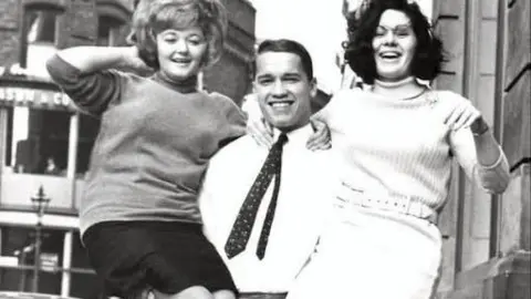 Roy Smyth/Belfast Telegraph A black and white archive photo of Arnold Schwarzenegger lifting two women into the air outside the Belfast Telegraph building in 1966. He has short, dark hair and is wearing a white shirt and a dark patterned tie. The women are wearing turtleneck jumpers and miniskirts. There are cars parked along the street in the background. 