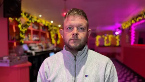 A picture of a man wearing a grey jumper. He has a short beard and short hair. He is stood in front of a bright pink bar with warm yellow lights.