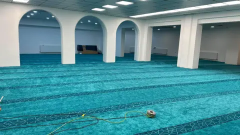 Salahaddin Community Centre The inside of the Salahaddin Community Centre mosque. The area shown is empty. Blue patterned carpets stretch from wall to wall. At one side of the room there are arches. There are lights in the ceiling.