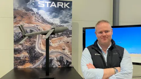 BBC Mike Armstrong - managing director of drone company Stark - looks at the camera, arms folded, wearing blue shirt and dark blue branded gilet. Next to him is a model of a drone and the Stark logo on a board with a drone shot of a coastal area.