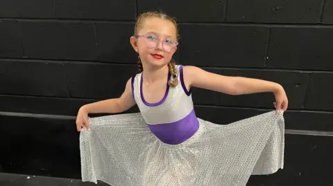 Leanne Rinne/BBC A young girl with glasses is doing a courtesy in a sparkling sliver and purple dress. She is looking at the camera, and behind her is a black painted brick wall.