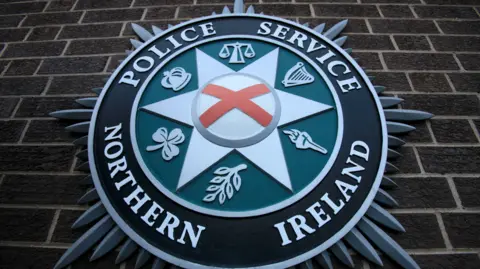 A close up of the PSNI emblem on a brick wall. It consists of a star shape in the centre, with the scales of justice, a harp, a torch, a leaf, a shamrock, and crown. 