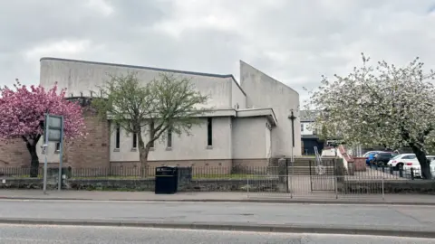 Modern-design grey church building with a pink cherry blossom tree on one side and a white cherry blossom tree on the other. It is at the side of a road and there are cars parked at the side.