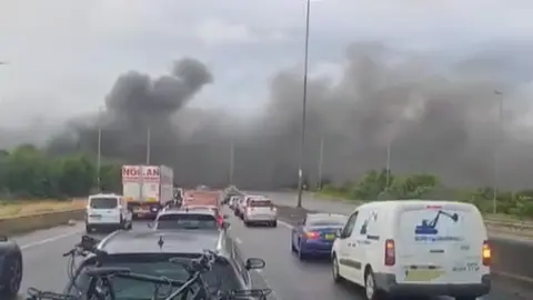 Tim Dixon A blurry photo of a busy motorway with thick black smoke billowing across both carriageways in the distance