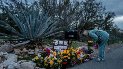A woman in a teal sweatsuit bends over and lays flowers at a memorial for Nancy Guthrie 