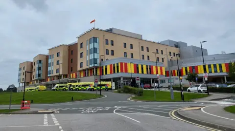 A hospital with ambulances parked outside it. The hospital building is several storeys high, and has red and yellow cladding on the lower part.
