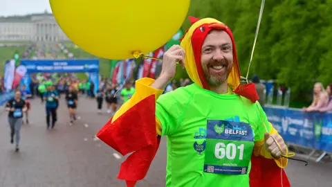 A man in a green t-shirt dressed in a chicken outfit at the start of the 2024 marathon. he's smiling, holding a yellow balloon and is running down Prince of Wales avenue at city hall 