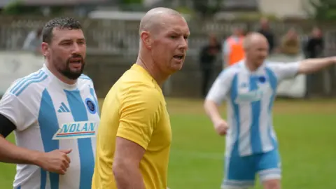 James Constable has a bald head and is wearing a yellow Oxford United strip. He is being marked by an Ardley player wearing a blue and white striped shirt.