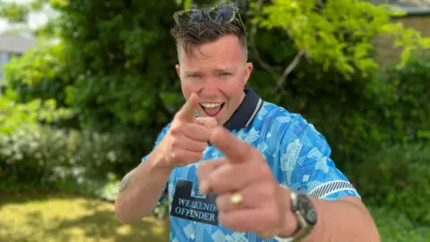 Sonny Green is pointing at the camera and smiling outdoors. He is wearing a blue top with a navy collar and has sunglasses on top of his brown hair.
