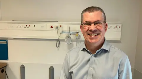 Amy Holmes/BBC A man wearing glasses and a light patterned shirt stands in a hospital treatment area, with wall-mounted medical equipment, outlets, and tubing visible behind. The setting is clean and clinical, with neutral walls, safety rails, and equipment panels indicating a patient care space under bright overhead lighting.