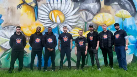 Eight people in a line stood in front of a blue and yellow mural. They are all wearing back t-shirts or hoodies with the words Hip Hop Healing on the front.