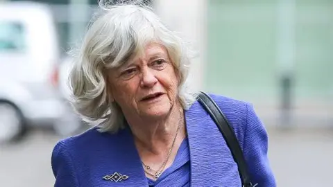 Getty Images Ann Widdecombe, seen in 2019 when she was elected as a Member of the European Parliament ( MEP) for South West England, wears a blue jacket and carries a shoulder bag while walking outdoors near a building. A parked vehicle and blurred background buildings can be seen behind her.