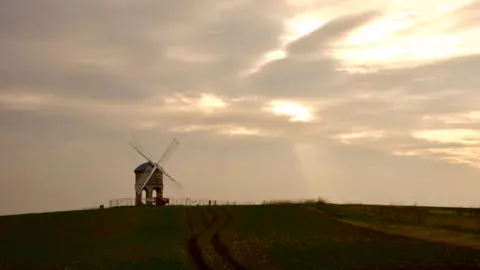 A round windmill with arches underneath it stands on a grassy hill on the horizon as the sun starts to set