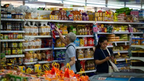 Getty Images An elderly Chinese man browses through shelves of packaged bread at a supermarket in Singapore, while a woman next to him checks her phone. On the shelves you can see different types of breads, packaged cakes, and jars of food.
