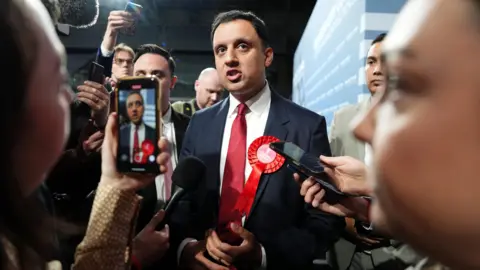 PA Media Anas Sarwar, in red tie with white shirt and dark suit, is surrounded by journalists with mics and phones. He is speaking.