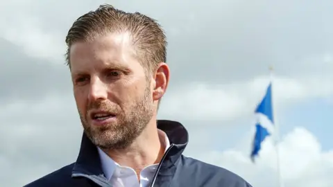 Eric Trump visible from the shoulders up. He is wearing a dark blue jacket and a saltire is visible in the background. He has swept back hair and a stubbly beard