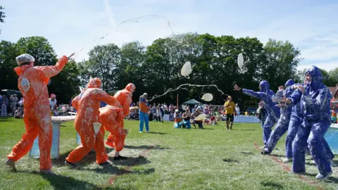 PA Media A group of people, standing opposite each other, each side wearing orange and purple. They are hurling custard pies at each other in a park.
