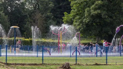 Several water fountains are spraying upward and outward from colorful structures. Children and a few adults are gathered inside the splash area, enjoying the water.
