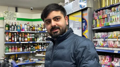 Kate Bradbrook/BBC A man wearing a blue coat which is zipped up. He has a beard and moustache. He is standing inside a shop with liquors and vaping supplies visible on the shelf behind him