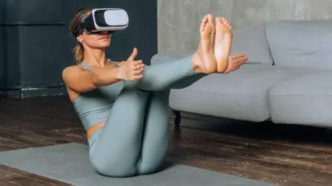 Getty image of woman pounding yoga pose on a mat in the living room while wearing VR headsets.