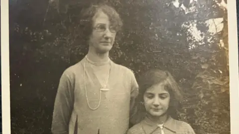 Marjorie Mitchell A grainy black and white photo showing Marjorie Mitchell as a 12 year old girl, with short dark hair, standing next to her mother who is wearing glasses and a pearl necklace. Marjorie is looking down and her mother is looking off-camera. 
