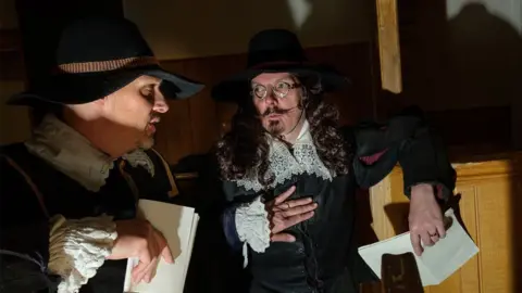 John Beardsworth Two men in mid-17th Century dress during a re-enactment of the Huntingdon witch trial. They are wearing black broad-brimmed hats and dark clothing from which white linen collars and cuffs can be seen, decorated with broad lace. They are holding white documents and appear to be conferring.